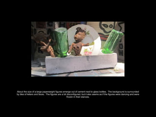 About the size of a large paperweight figures emerge out of cement next to glass bottles. The background is surrounded
by tiles of letters and faces. The figures are a bit disconfigured, but it also seems as if the figures were dancing and were
frozen in their stances.
 