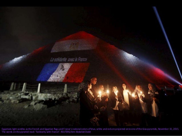 People stand in front of the Brandenburg gate which is illuminated in blue, white and red in the colors of the French flag...